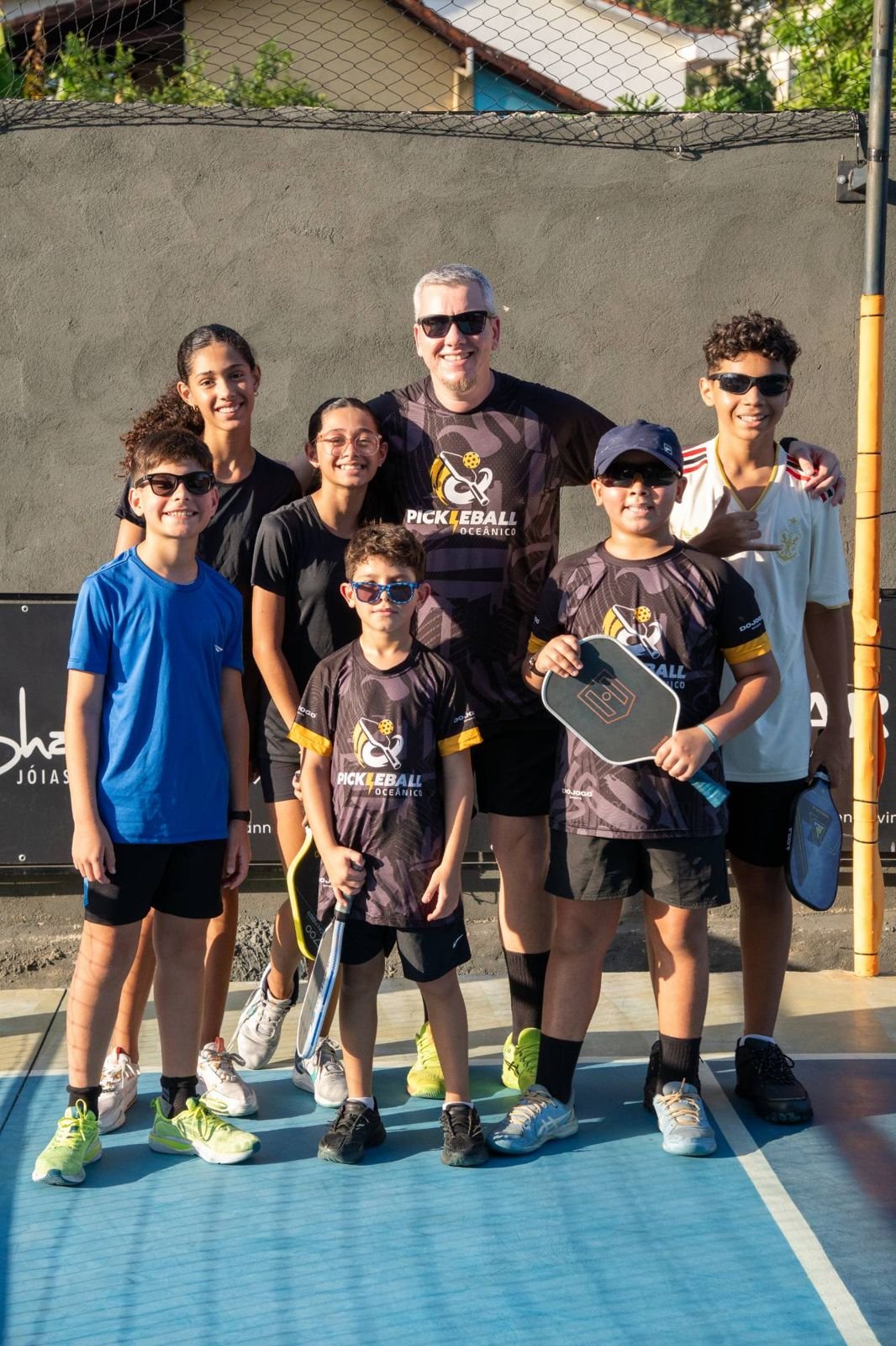 Professor e alunos da turma kids sorrindo na quadra com as raquetes